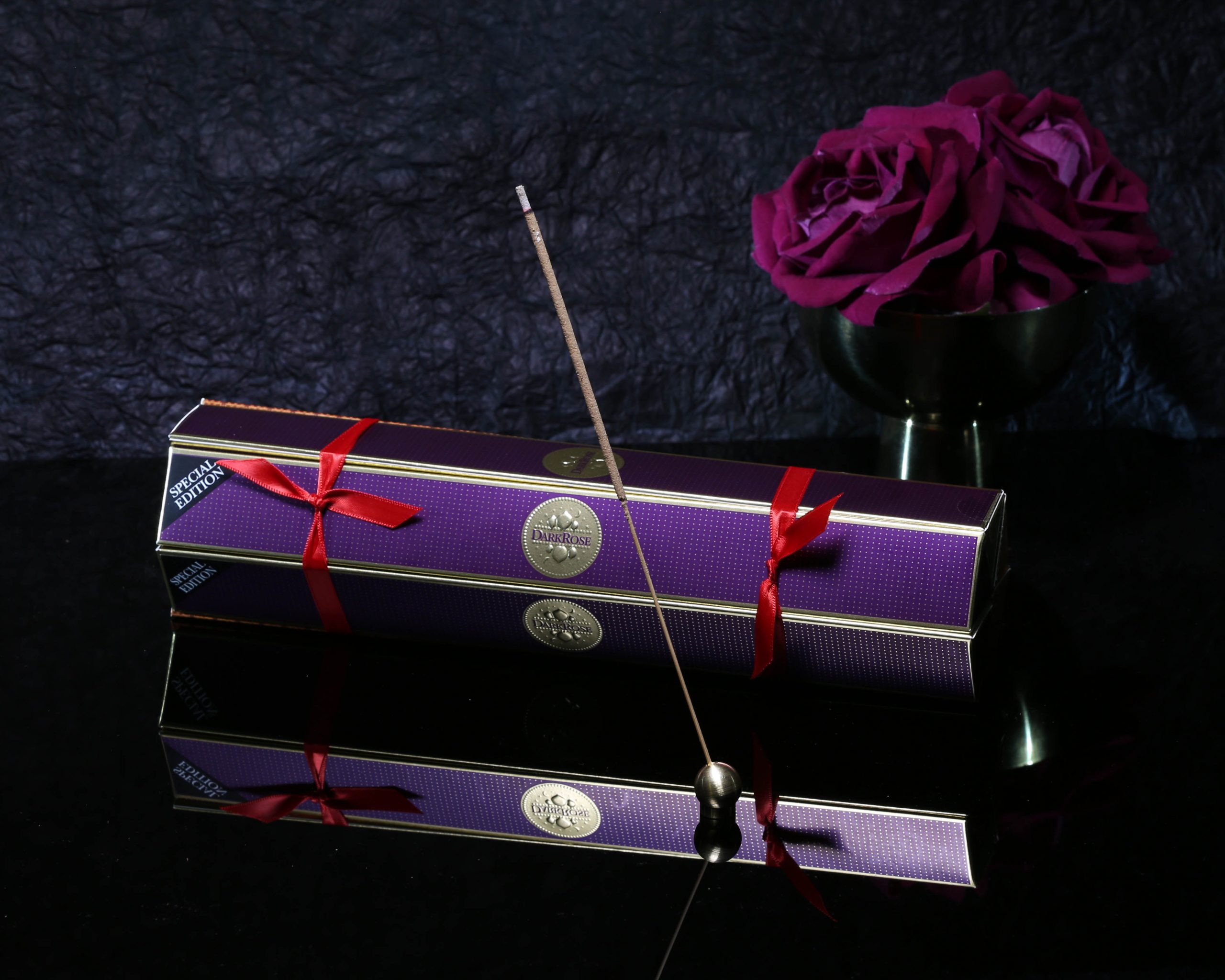 Czech & Speake Dark Rose Incense Sticks bundle pack on shiny black surface and dark background with rich pink roses behind and incense stick in holder in front.