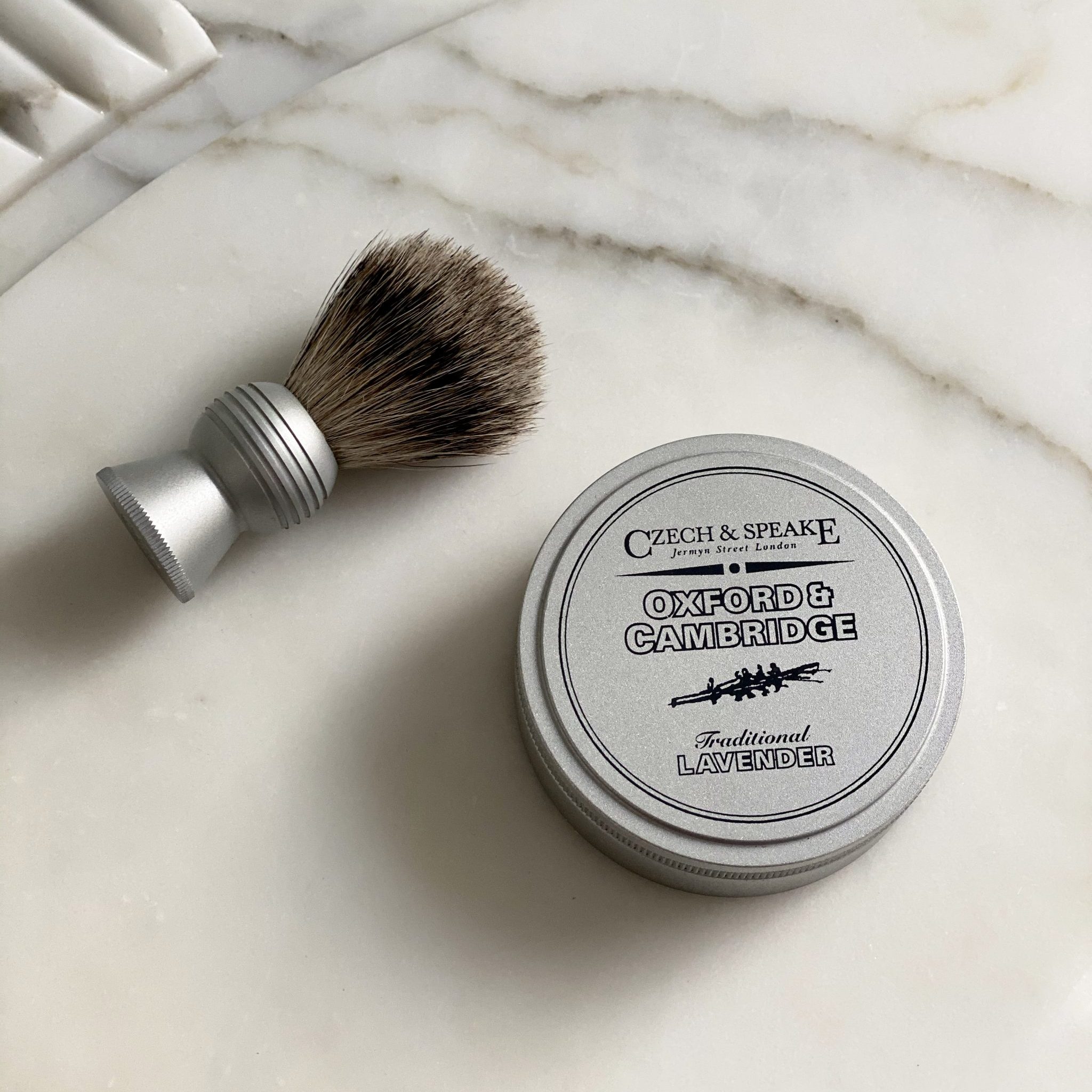 The Oxford & Cambridge Travel Shaving Brush and Shaving Soap Dish lie on a marble bathroom sink surface